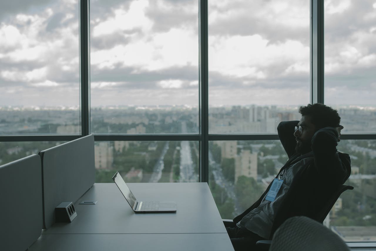 A man sits in an office with hands on head in front of a laptop, overlooking a cityscape.